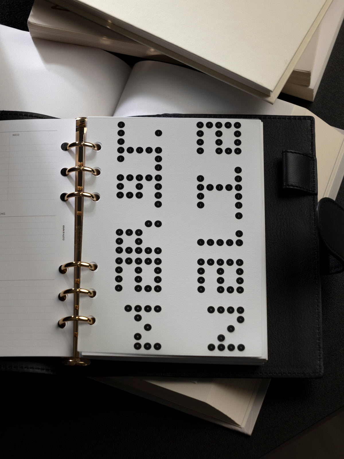 Six ring planner open on a black leather cover, showing a Scantron dashboard with bubbles filled to form letters, held by gold rings, with a lined insert facing and notebooks stacked behind.