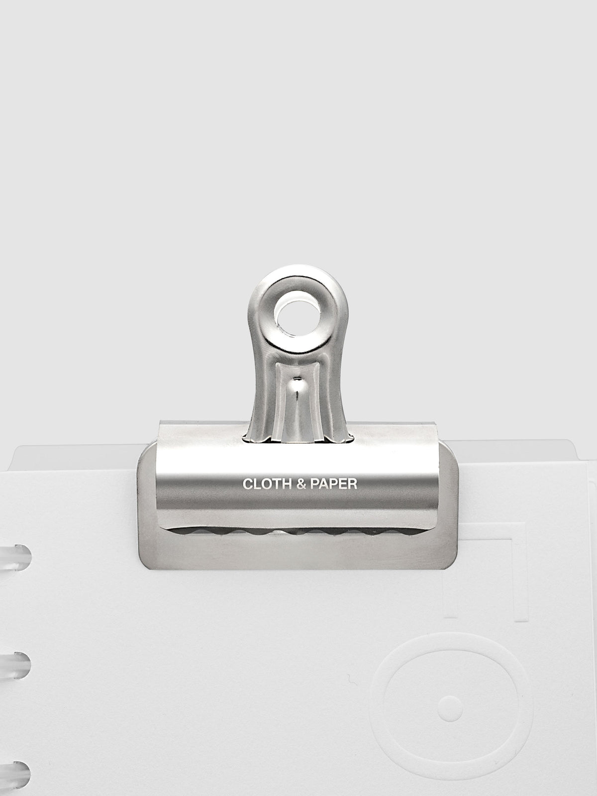 Silver metal notebook clip with Cloth & Paper logo etched onto the surface shown clipped onto a notebook.