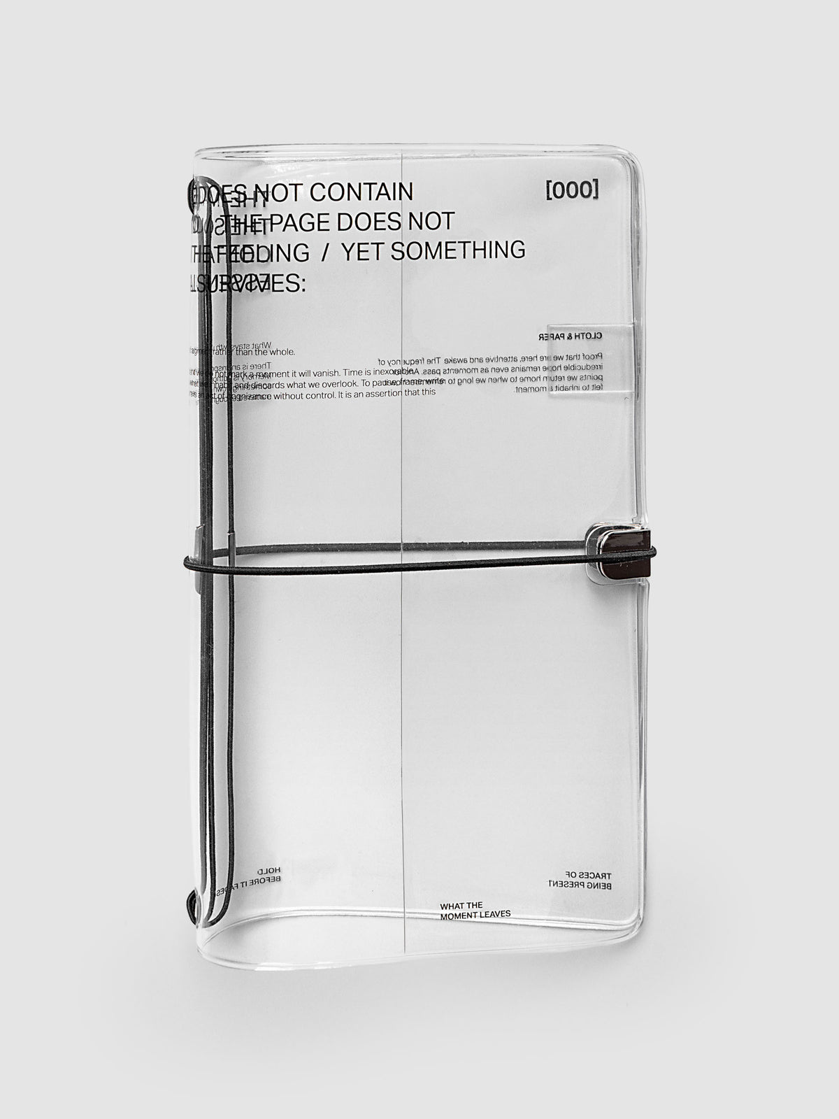 What Lasts Travel Notebook Cover in clear vinyl, A5 Slim, front exterior view, closed and upright against a light gray background, showing black typographic text printed on the vinyl and a black bungee cord with silver metal closure