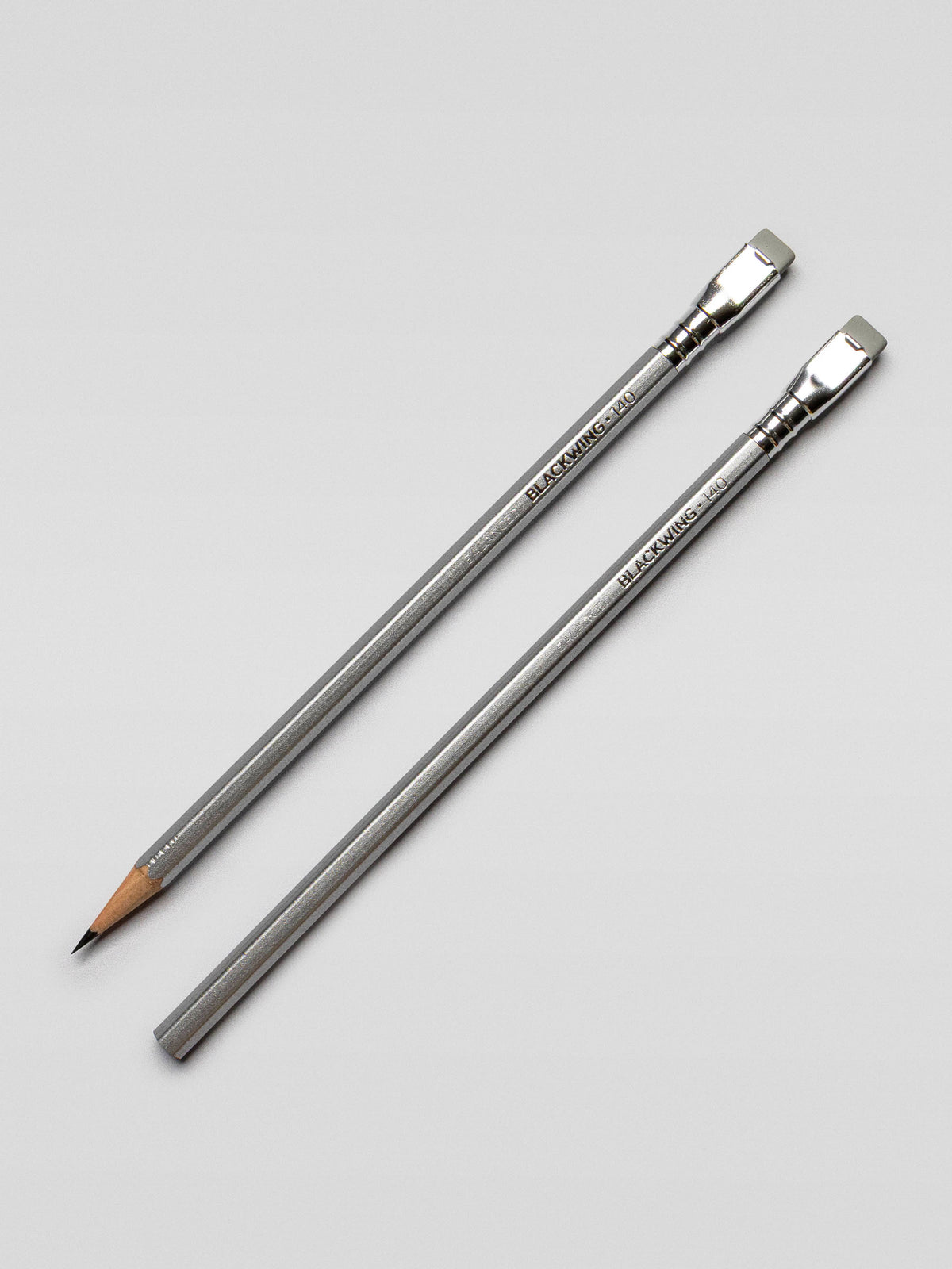 Two Blackwing 140 pencils laid side by side on a light surface, one sharpened and one unsharpened, highlighting the metallic finish.