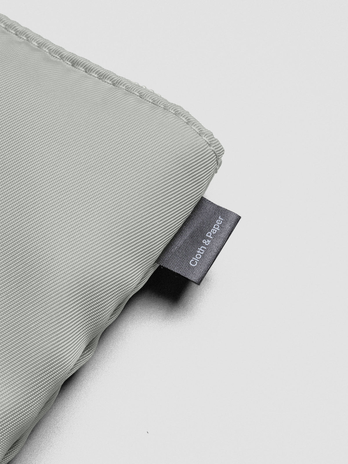Close up of the stitching and tag on the green-grey nylon, padded bag reading "Cloth & Paper".