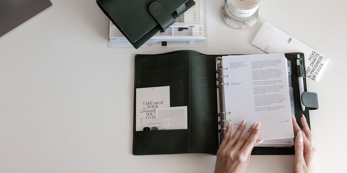 A Foundations Leather 6-Ring Agenda in A5 is opened on a white desk. Two hands are beginning to flip through the pages inside. Above is a Foundations Leather 6-Ring Agenda in Personal with other surrounding planner accessories.
