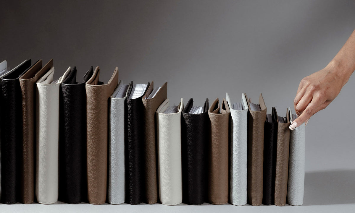 An image representing folios from our new Heirloom Leather Collection. A hand is reaching for a Folio in color Veleta. The different folios and colors are standing up side by side with their spines facing out.