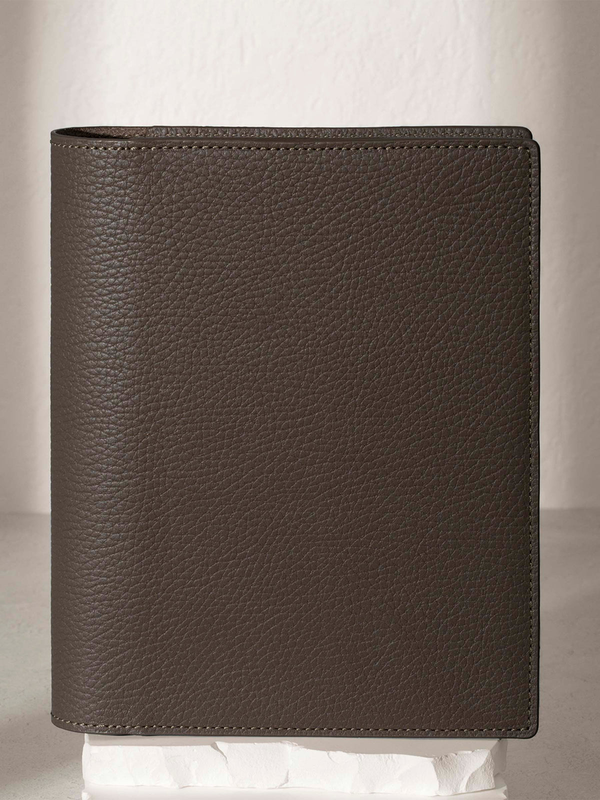 Small Leon folio displayed on a white stone pedestal. The background is a natural textured off-white material, and a spotlight behind the folio highlights its placement.
