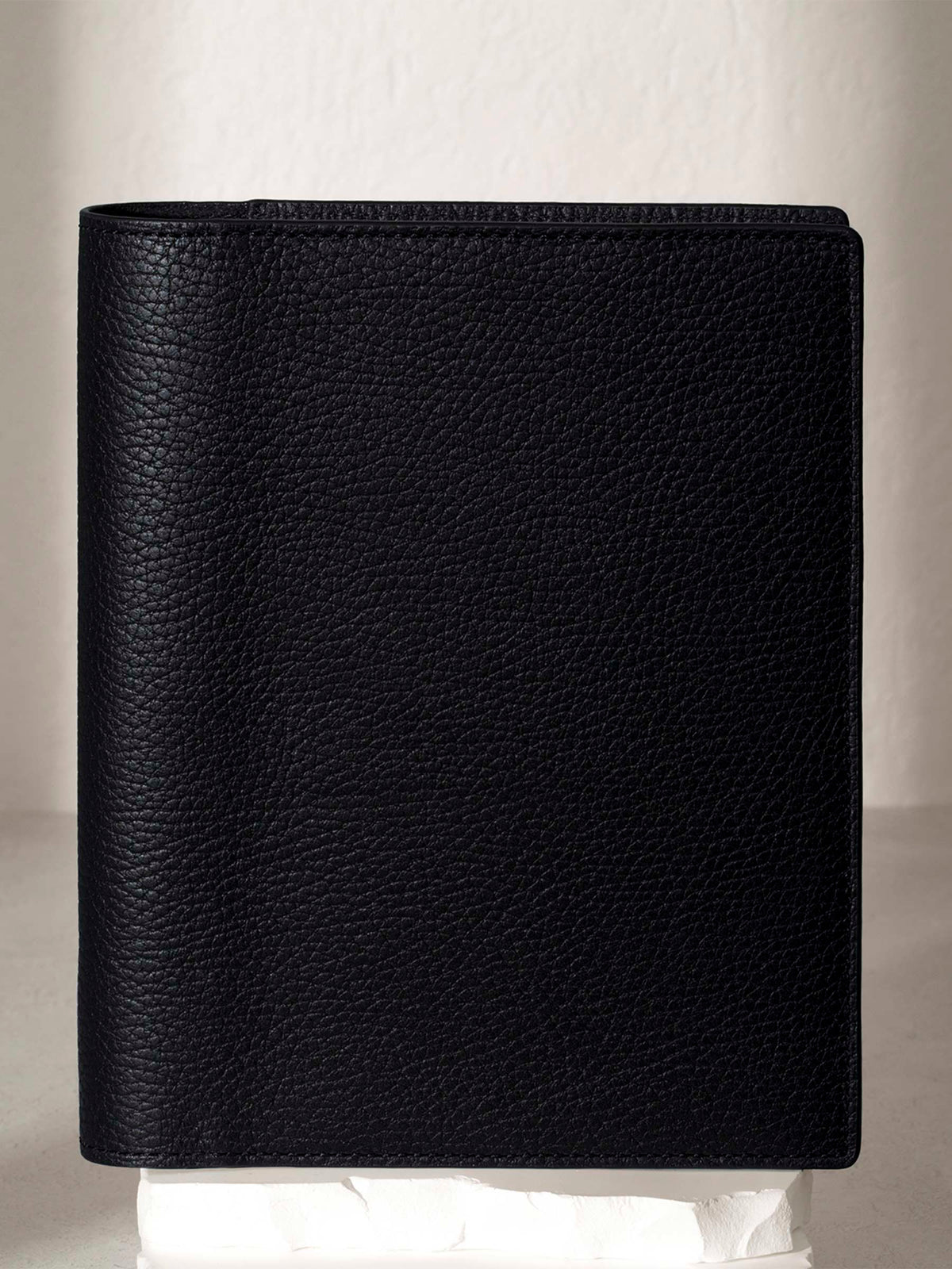 Heirloom Leather Folio, Small, Cloth and Paper. Padrillo folio displayed on a white stone pedestal. The background is a natural textured off-white material, and a spotlight behind the folio highlights its placement.
