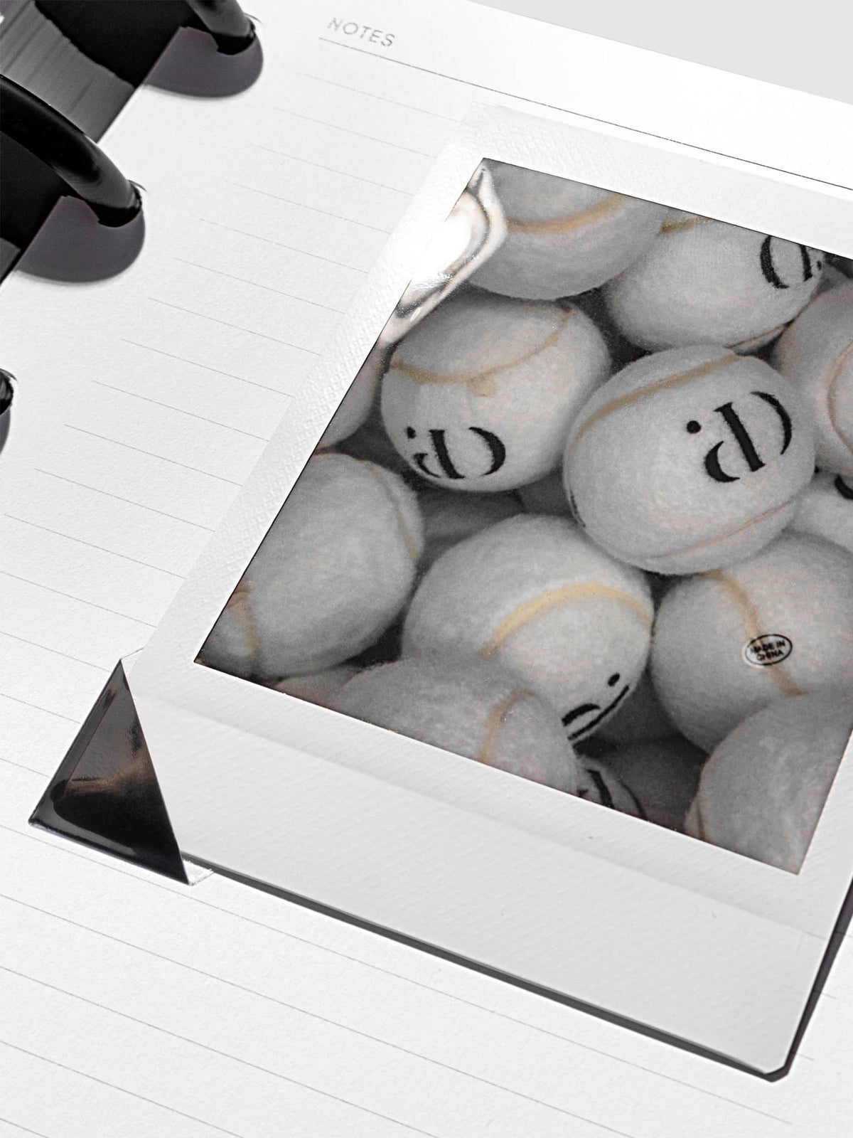 Black photo corner stickers in use, shown holding a photograph of tennis balls inside a notebook.
