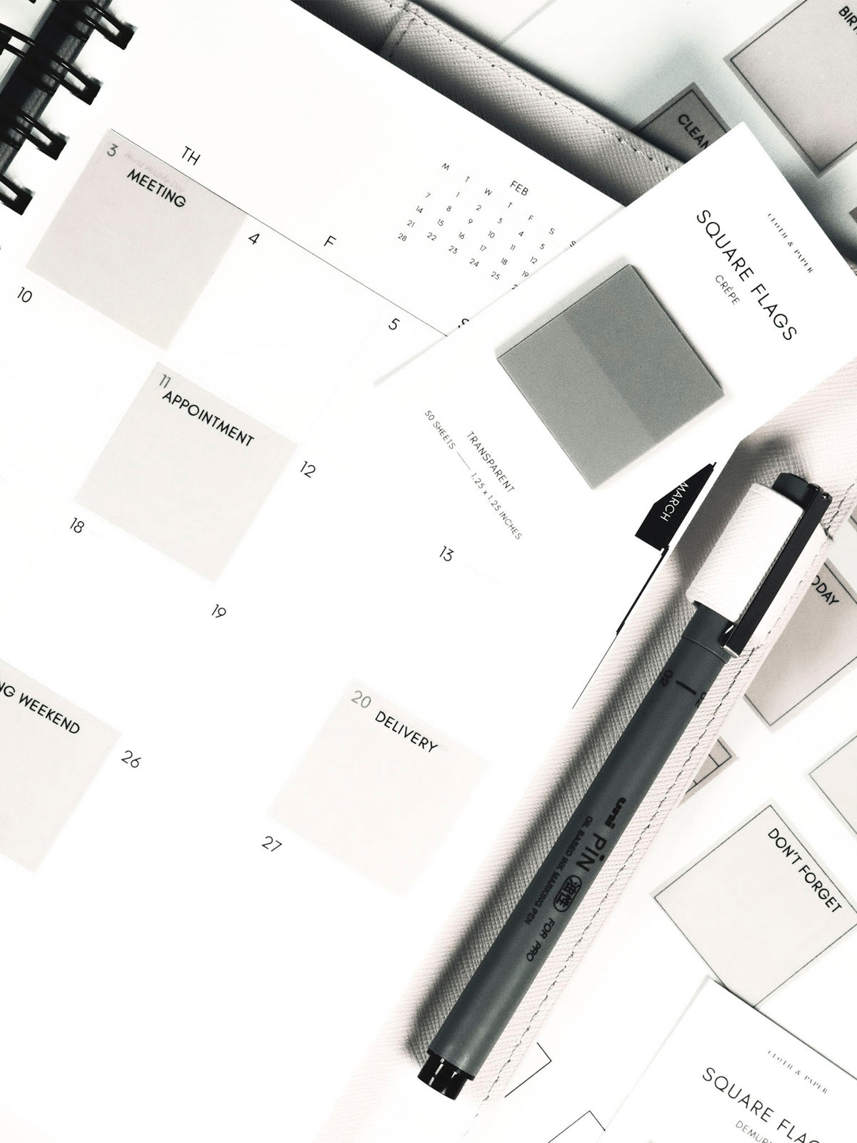 Square Page Flag Printable | Essentials page layered under a planner showing page flags in use. A Uni Pin Marking Pen is in the planner's pen loop and a set of square page flags are resting on top of the planner.