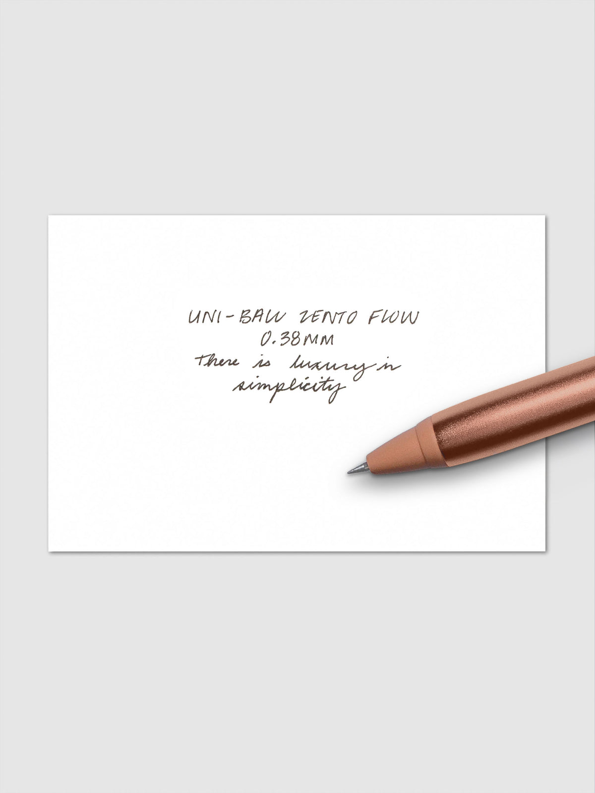 Handwritten writing sample using the Uni-ball Zento Flow Pen in a 0.38 mm nib. Shown with copper colored pen resting across paper.