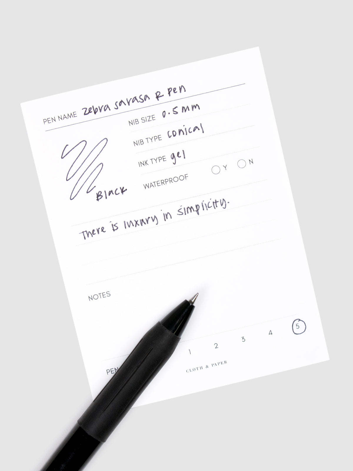 Zebra Sarasa R Pen, 0.5 mm, Black Ink, Black Body, Cloth and Paper. Pen resting on pen test sheet displaying writing sample.