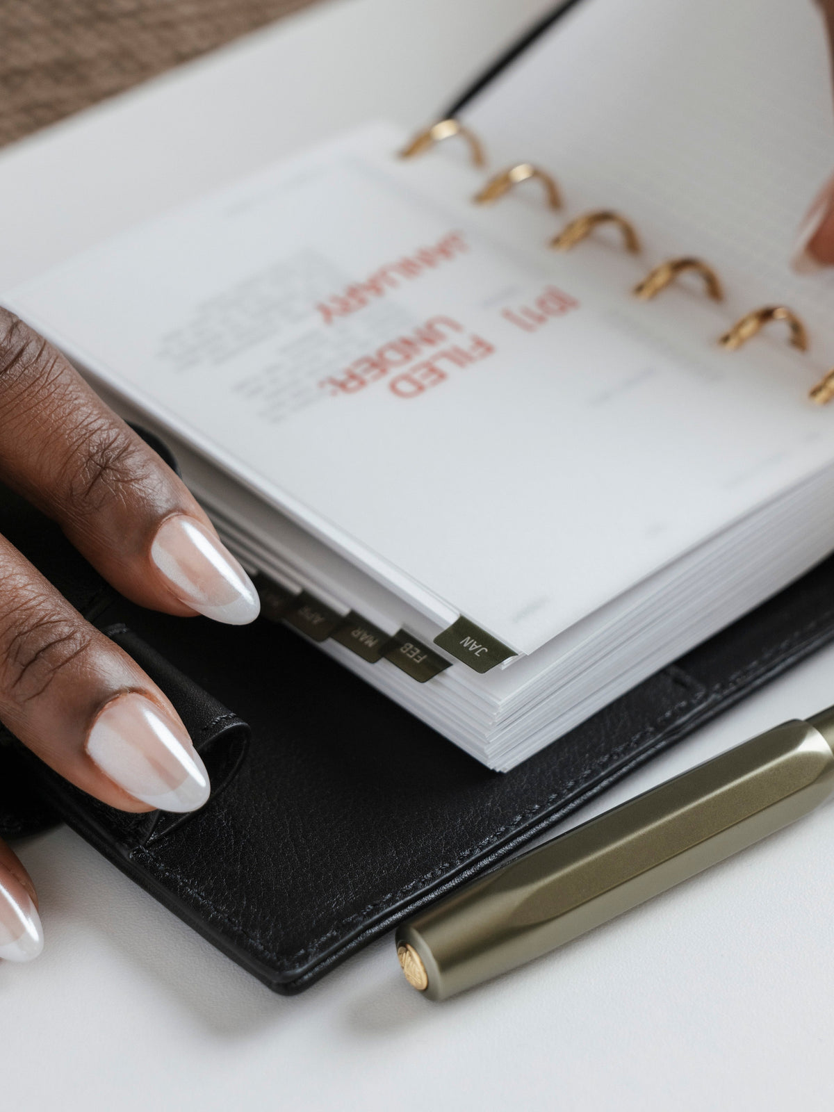 A close-up of a hand with long, white almond-shaped nails holding open a black leather ring bound planner.