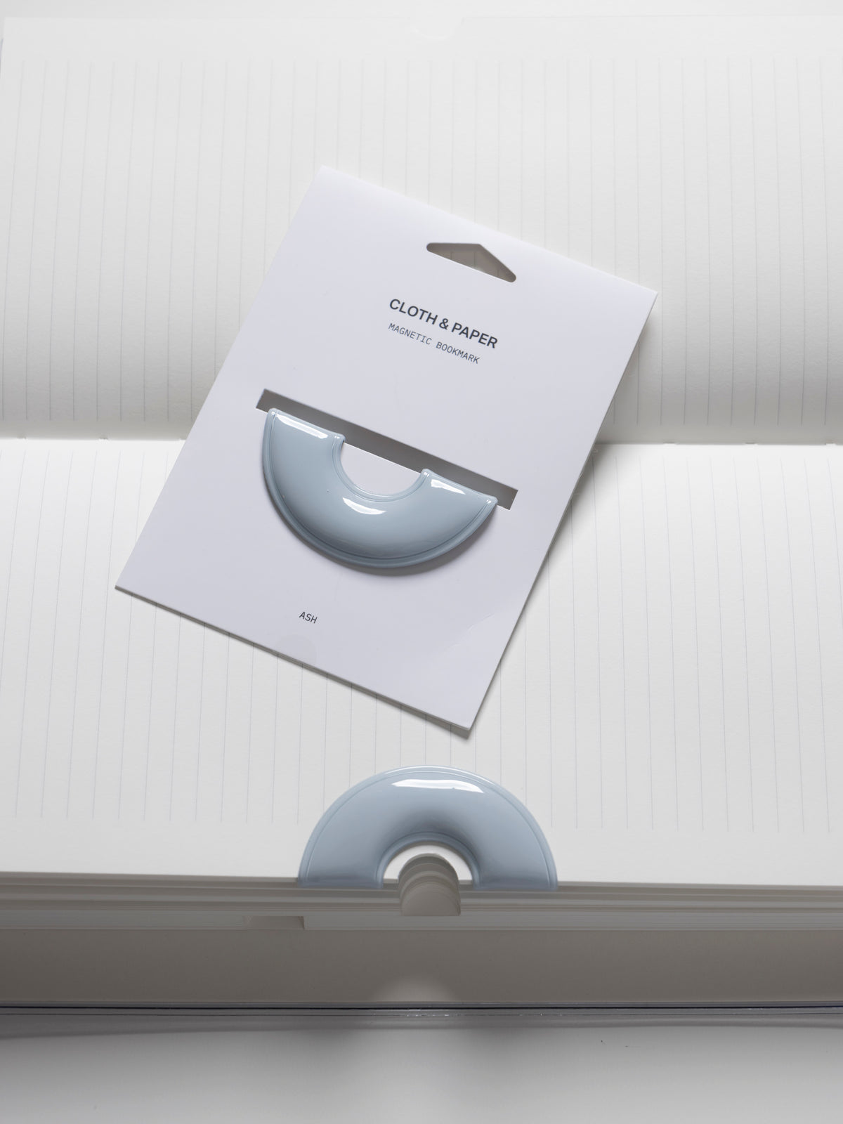 Magnetic Bookmark | Spotlight in the color Ash, a light grey, shown on a backing card with one in use in a notebook below