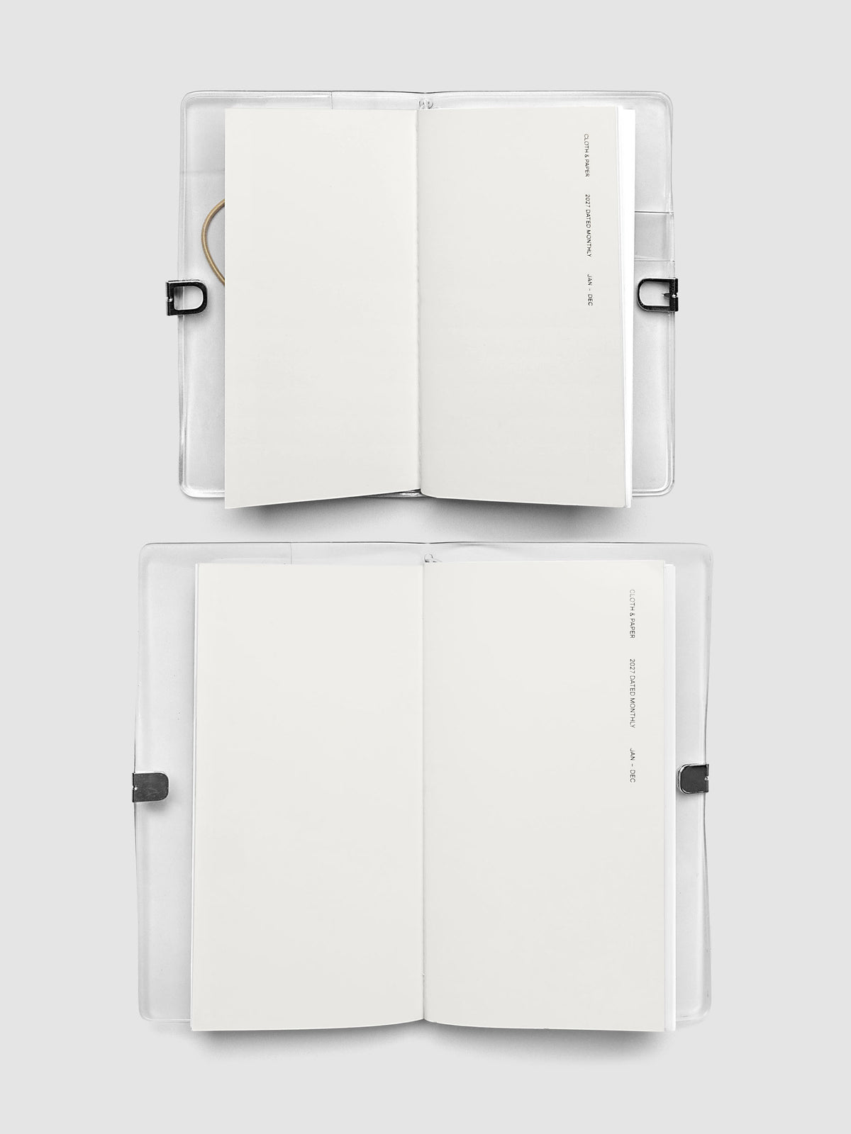 Two open travel notebooks stacked in their clear vinyl covers, both showing a blank interior page and 2027 Dated Monthly January through December cover text, comparing Pocket Plus and A5 Slim sizes