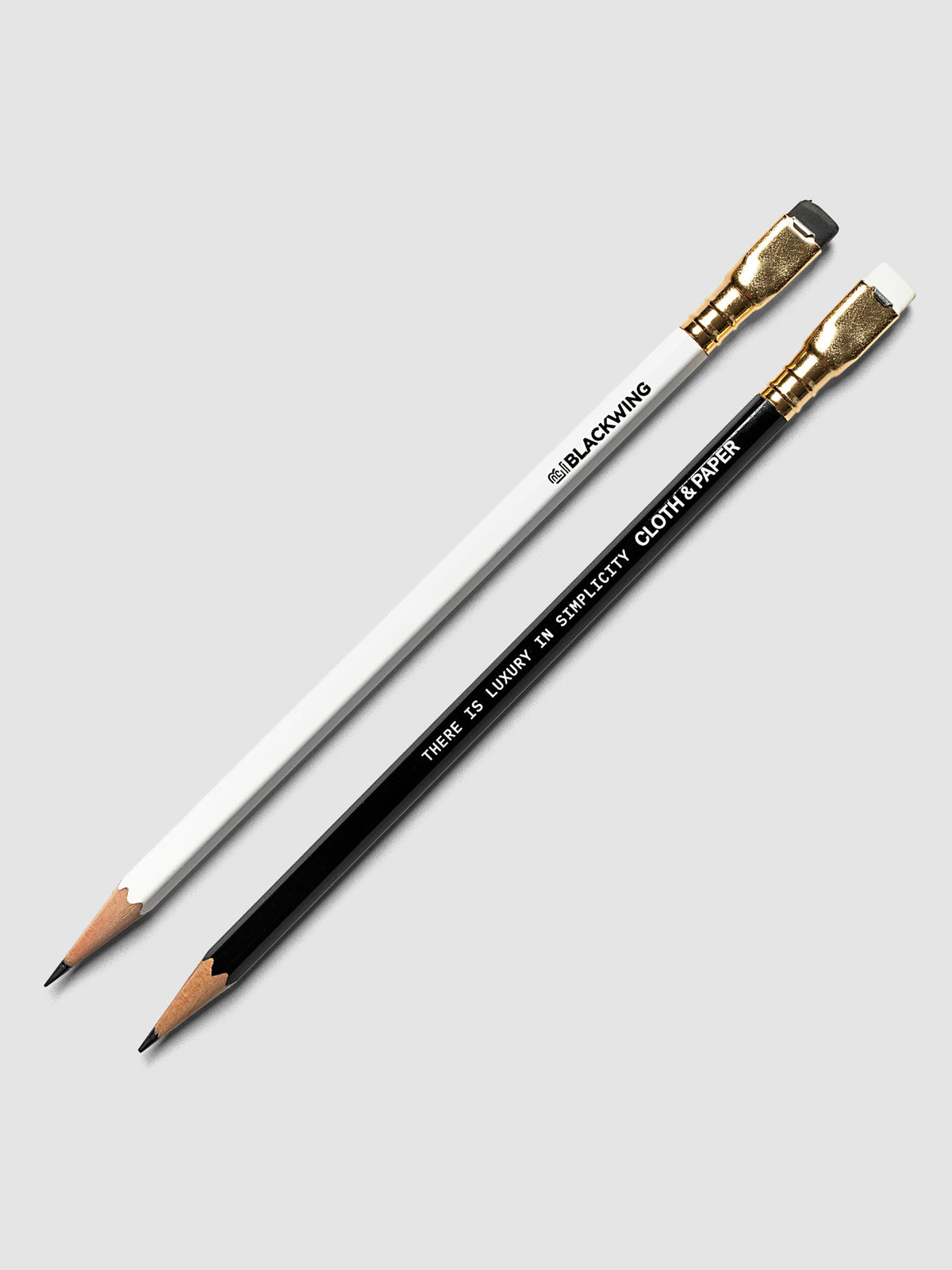 Two pencils, one white and one black, both with gold ferrules and erasers, laid diagonally on a light gray background. The white pencil is branded 'BLACKWING', and the black pencil reads 'THERE IS LUXURY IN SIMPLICITY' with the Cloth and Paper logo. 