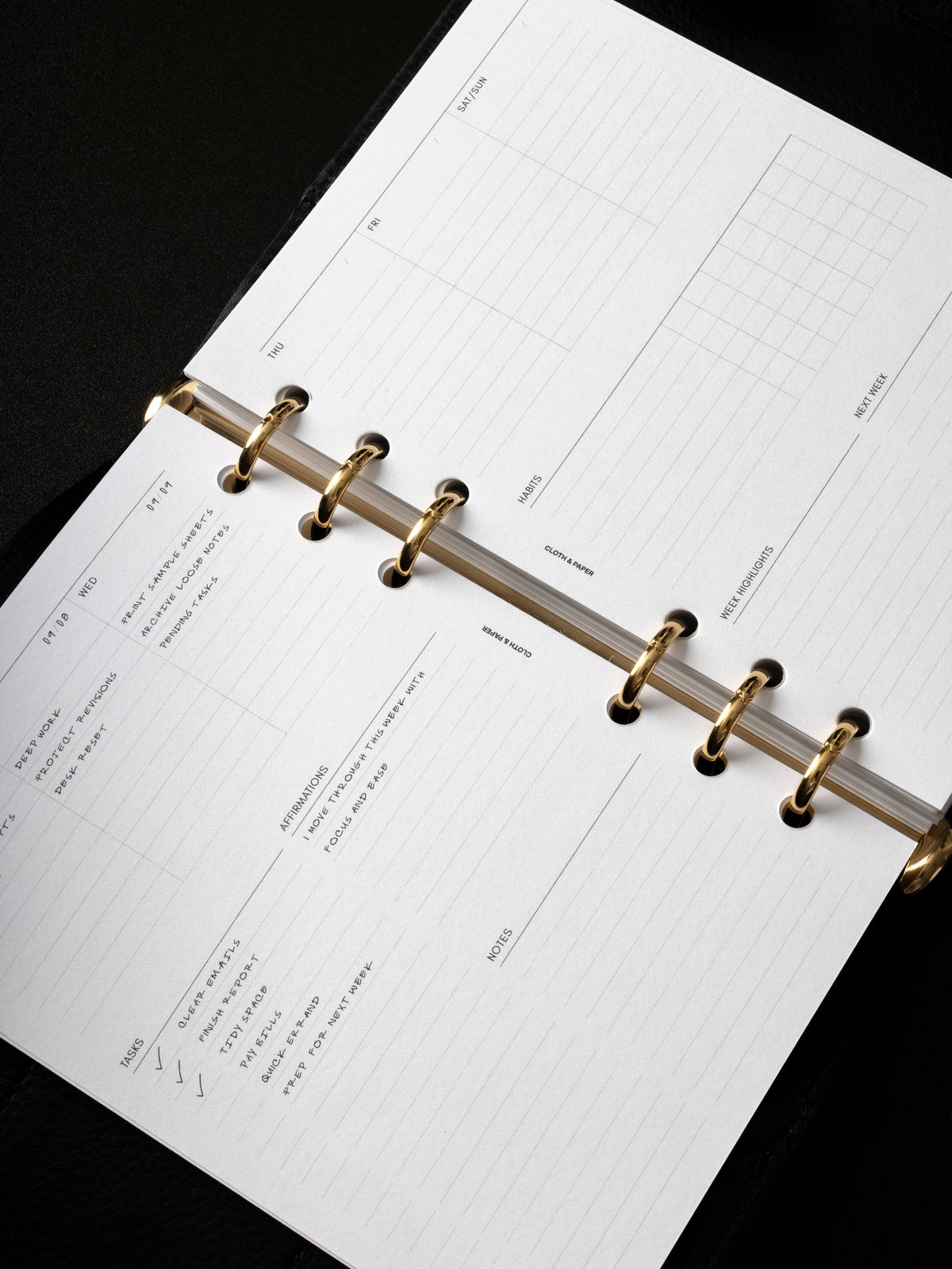 Six ring planner open at an angle to the Weekly Essentials insert filled in by hand, with gold rings and columns for tasks, affirmations, habits, notes, and week highlights.