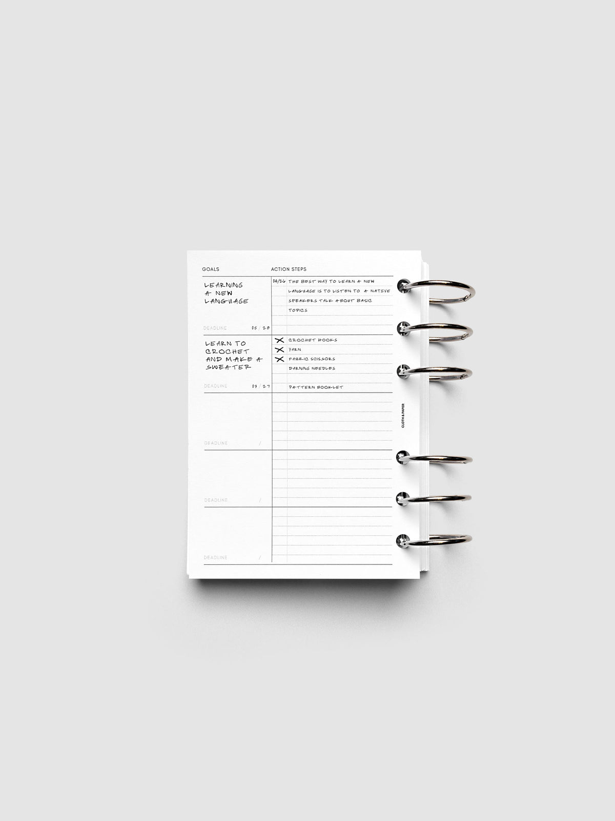 Open ring-bound planner displaying a goals and action steps page with sections for cleaning, language, and tactical project tasks. The planner is placed on a plain light gray background.
