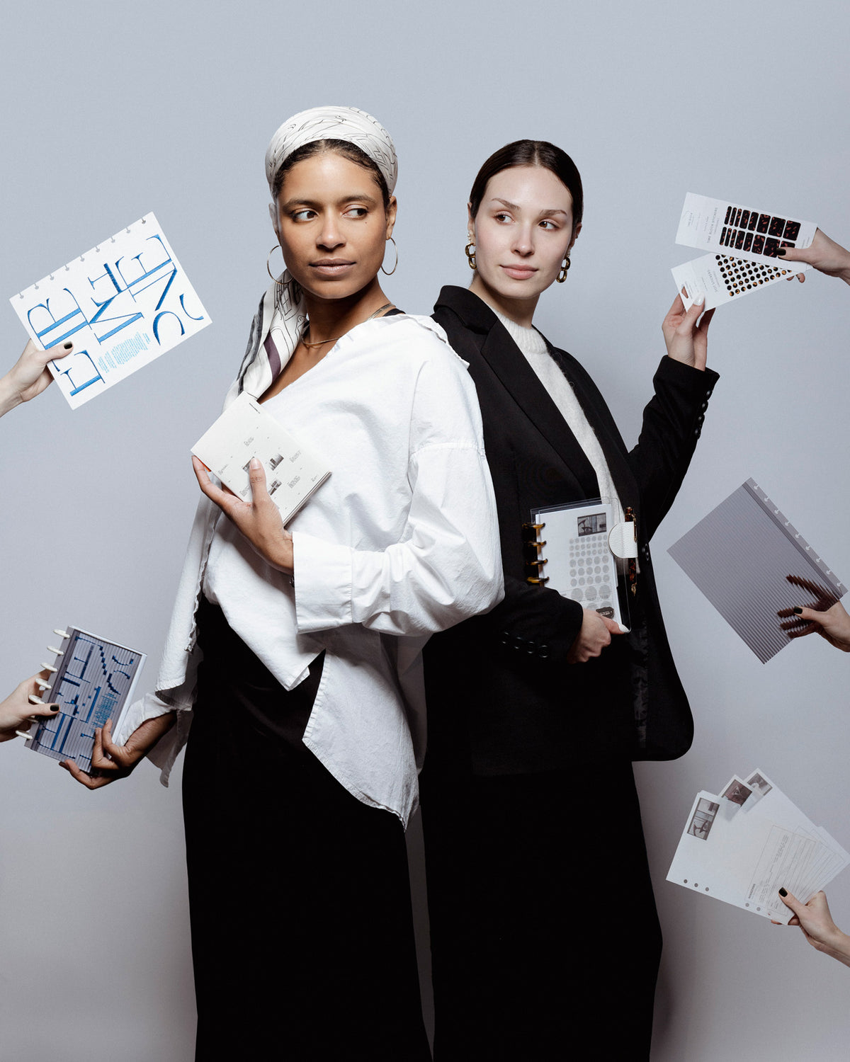 Two models are standing against a light gray background holding planner items from the Front Row Collection as hands coming in from the left and right are attempting to hand them more of these planner essentials.