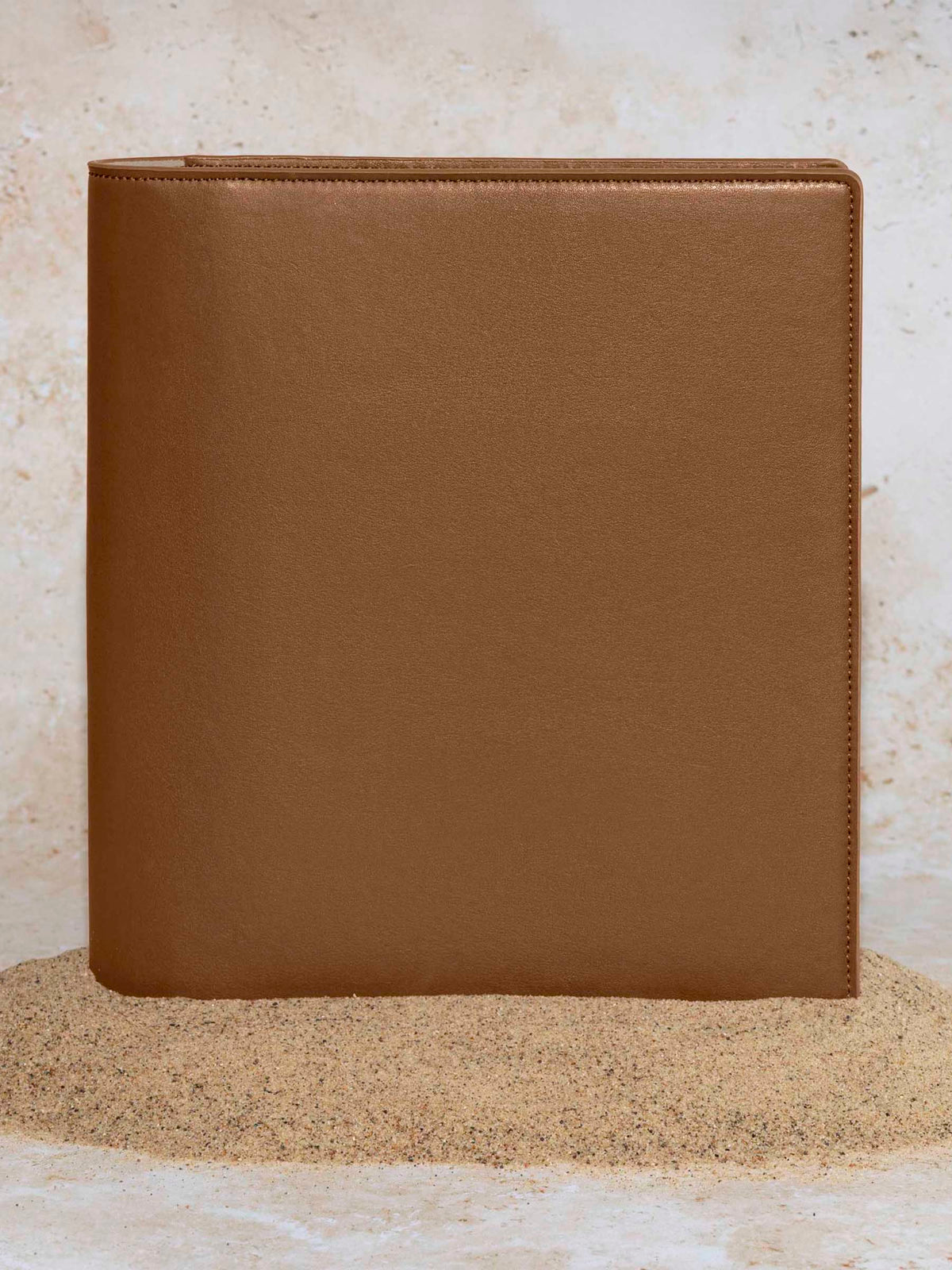 Folio displayed on a textured tan background, placed in a pile of sand. Color pictured is clay brown. 