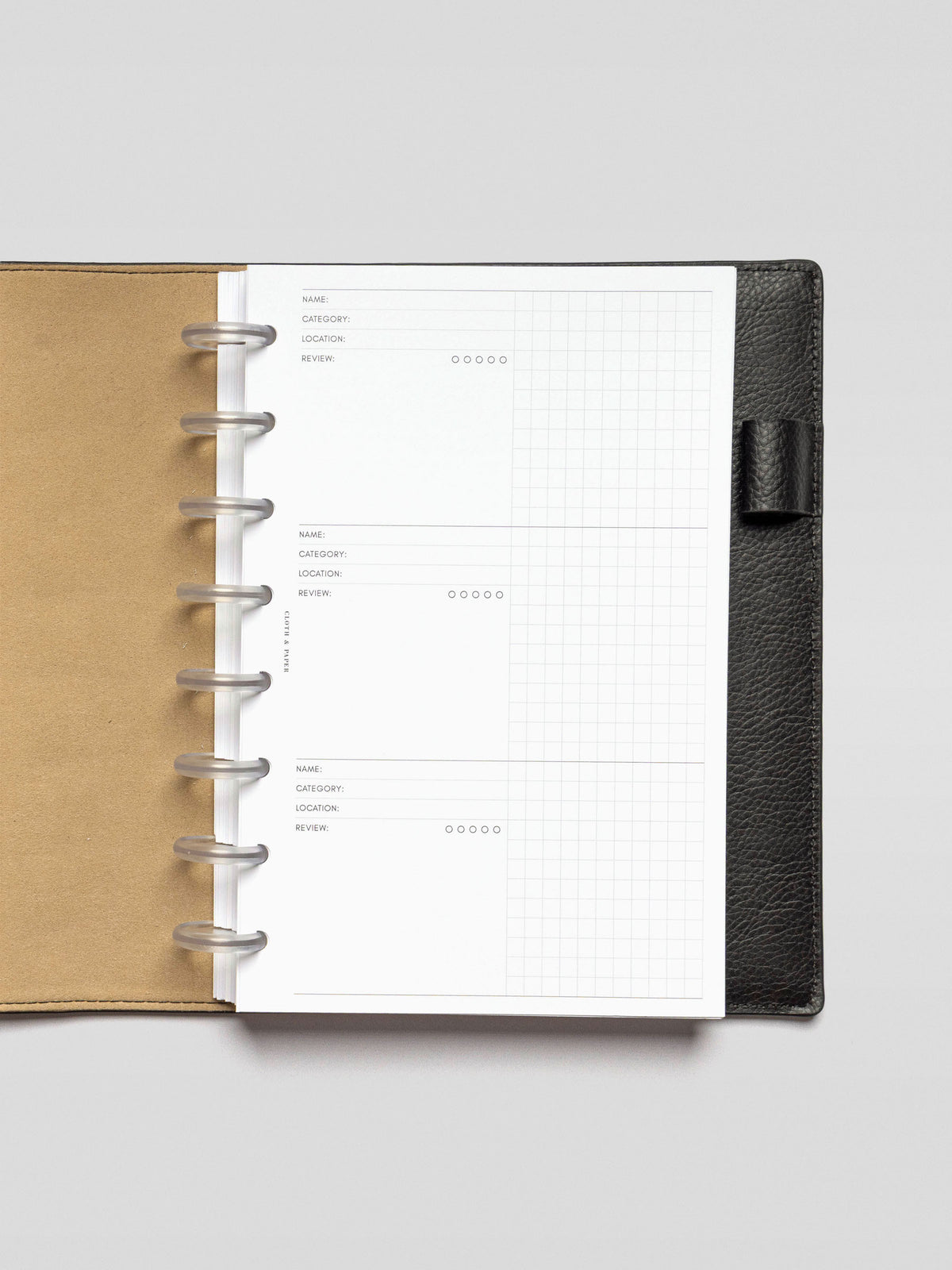Overhead view of a Half Letter size Experience Tracker Insert in a black leather planner with frosted discs. The insert features three structured sections for logging experiences, each with fields for name, category, location, review, and a five-dot rating system. A subtle grid appears on the right side for additional notes or journaling.
