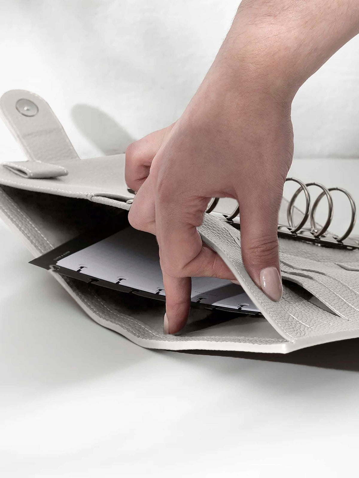 A fair-skinned hand shown opening the back pocket of the ash leather agenda on a white background. There are inserts and a dashboard inside the pocket.