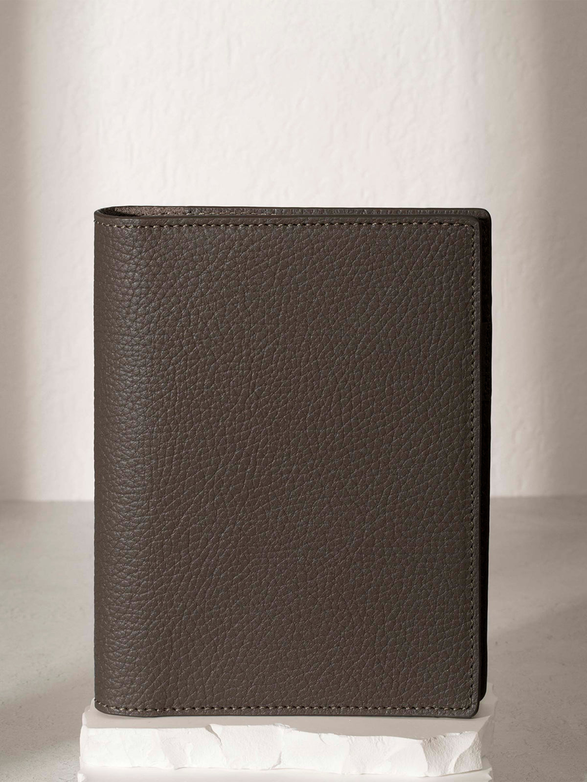 CP Petite Leon folio displayed on a white stone pedestal. The background is a natural textured off-white material, and a spotlight behind the folio highlights its placement.