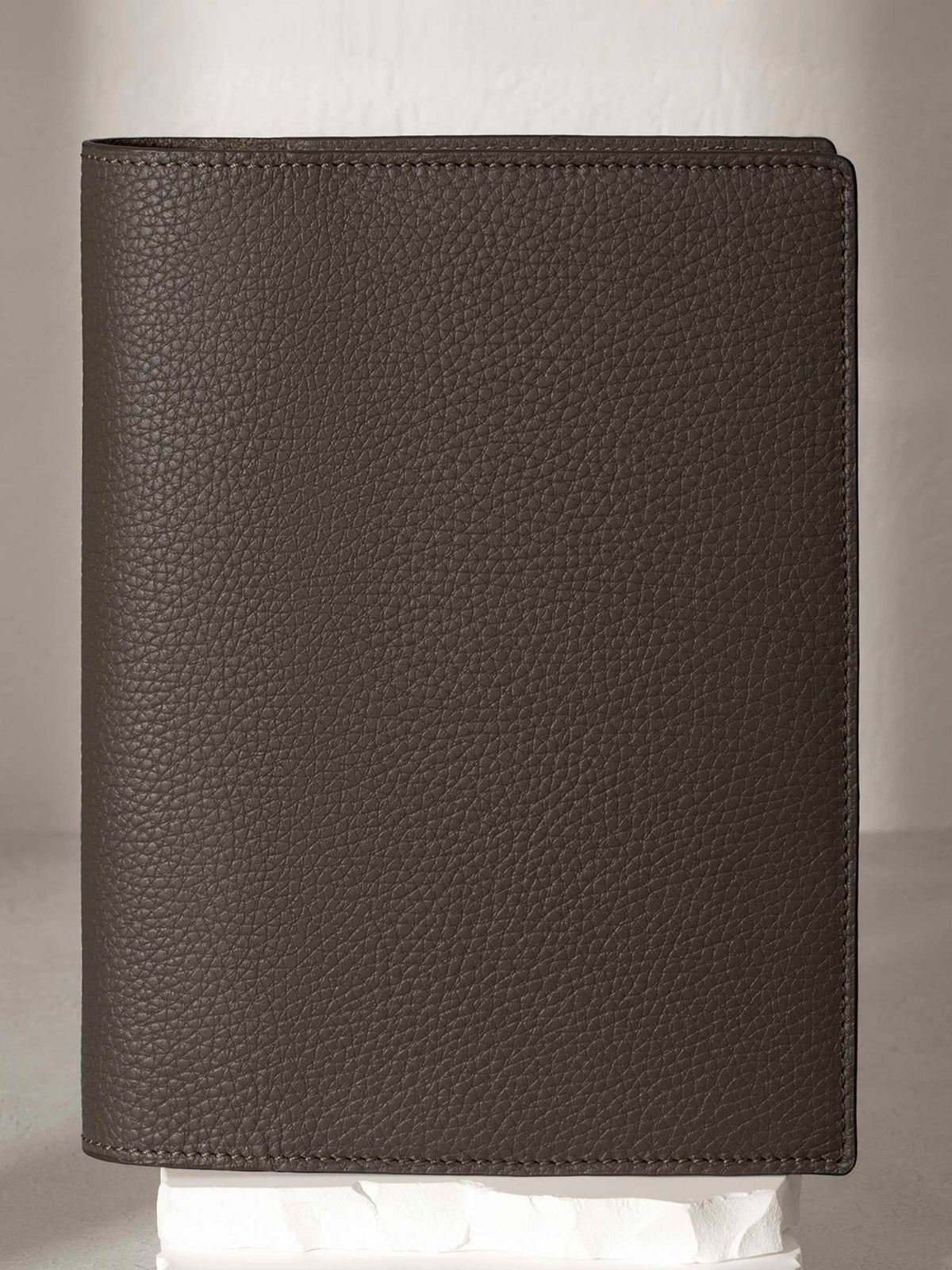 Large Leon folio displayed on a white stone pedestal. The background is a natural textured off-white material, and a spotlight behind the folio highlights its placement.
