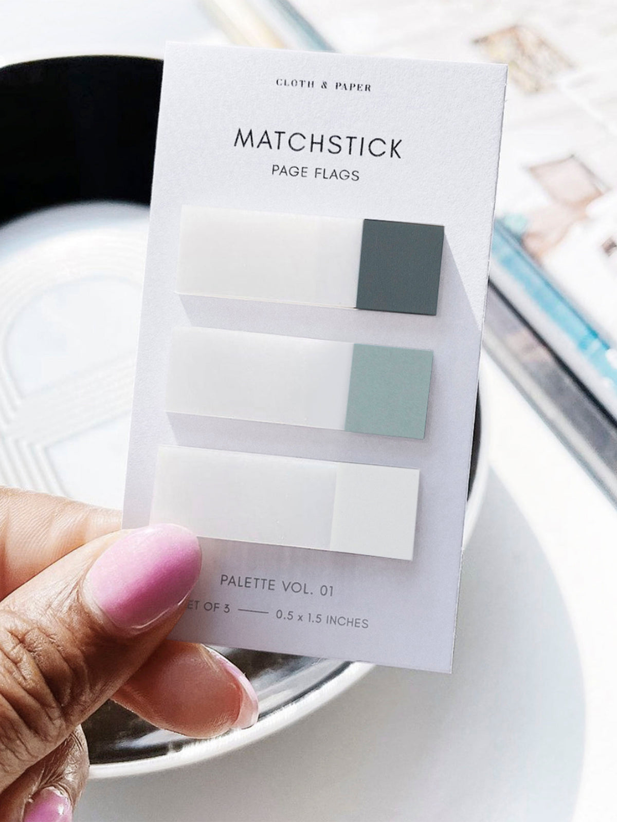Matchstick Page Flag Set | Palette Vol. 01 | Lagoon, Mykonos + Aspen | Cloth and Paper. Photo of page flags on their backing being held in the center of the frame. There is a decorative bowl and magazine in the background.