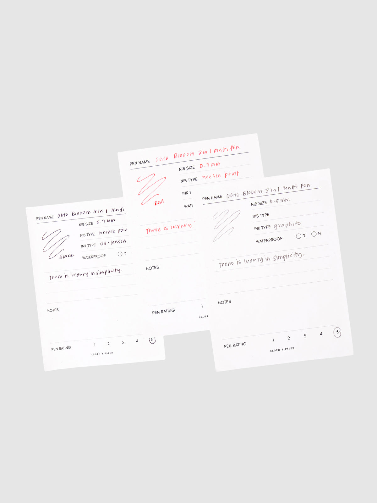 Ohto Bloom 3 in 1 Multi Pen, Cloth and Paper. 3 pen test sheets with writing samples for black ink, red ink, and mechanical pencil layered on top of each other against a neutral background.