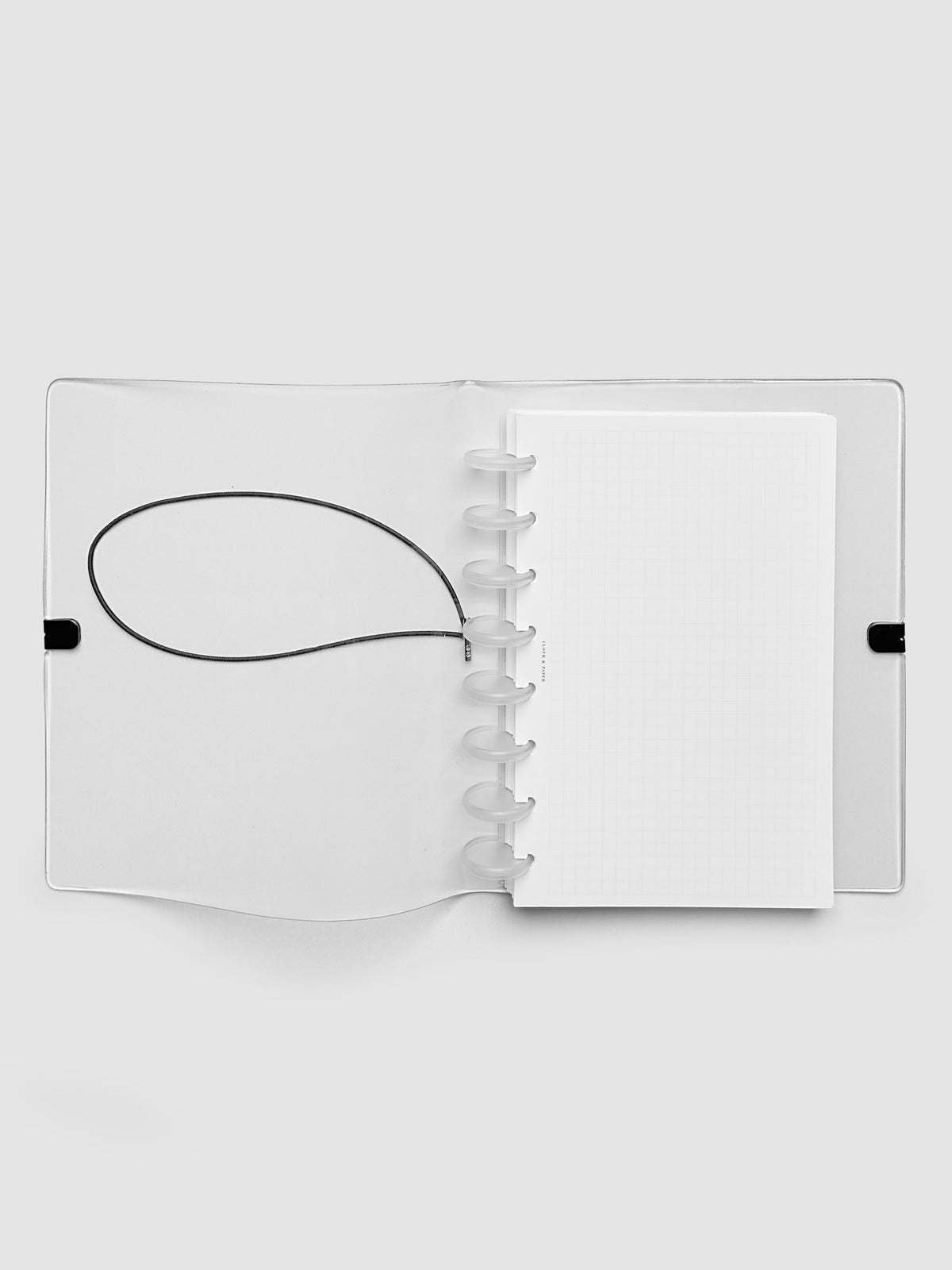 An opened clear plastic notebook cover with a black elastic closure band, with a discbound planner and grid inserts inside, against a light gray background.